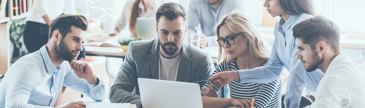 Um grupo de jovens colaboradores reunidos na mesa do escritório, engajados em trabalho e comunicação, enquanto seus colegas estão ocupados ao fundo.