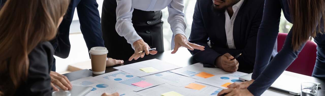 Nessa imagem, é possível observar uma sala de reunião com colaboradores diversos ao redor de uma mesa corporativa. Na superfície da mesa, há relatórios, gráficos de análise e post-it’s. Aparentemente, uma ferramenta de agile está sendo ensinada.