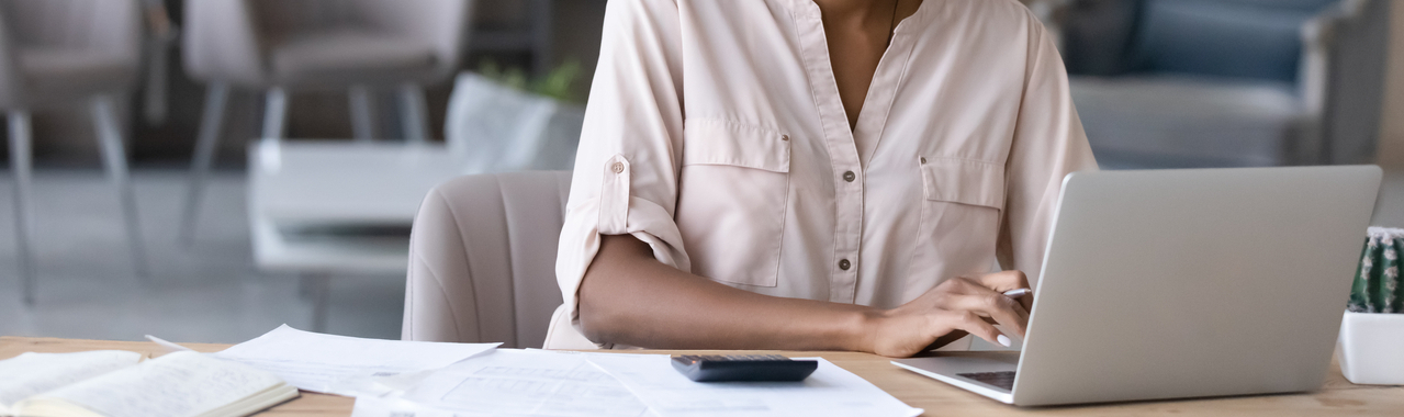 Na imagem, vemos uma pessoa sentada em frente a uma mesa, com um notebook aberto à sua frente. Ela está digitando algo, enquanto segura uma caneta na mão, e com alguns papéis espalhados pela mesa. À esquerda do notebook, há uma calculadora, indicando que ela está realizando cálculos, simbolizando o ato de calcular o LTV.