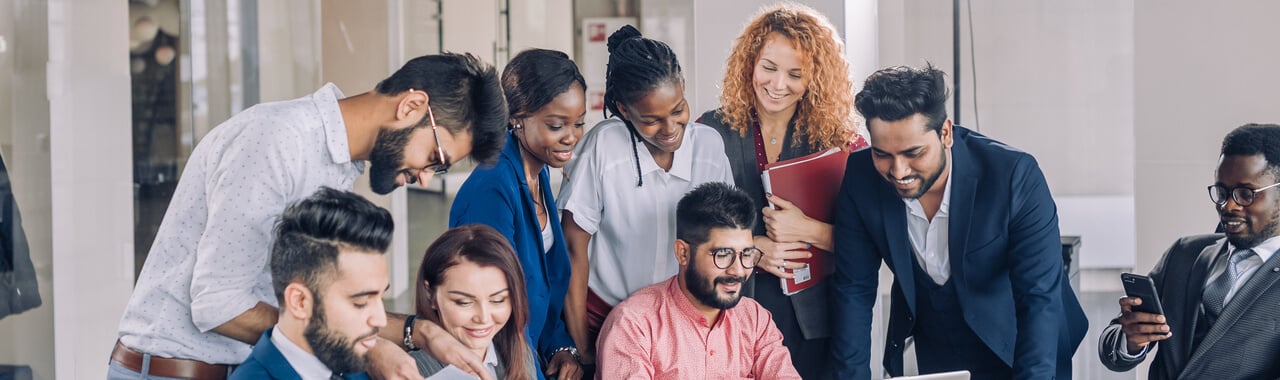 Num escritório corporativo, nove pessoas diversas interagem com alegria. No primeiro grupo, dois homens com traços do sul asiático e uma mulher branca concentram-se em um papel. Ao lado, o segundo grupo - composto por duas mulheres afrodescendentes, uma mulher ruiva e dois homens com traços do sul asiático - colaboram em um notebook. Próximo a eles, um homem afrodescendente utiliza o celular. A diversidade é evidente e a sinergia entre eles é palpável.