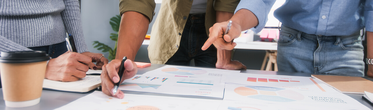 A imagem mostra uma mesa de trabalho com papéis que contêm gráficos e informações, rodeada por três pessoas que estão gesticulando e apontando para os documentos;eles estão trabalhando juntos para criar e implementar estratégias de negócios bem planejadas, representando o conceito de planejamento estratégico de TI.