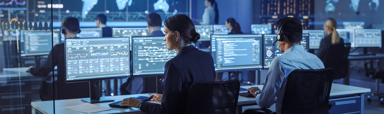 Imagem com um grupo de engenheiros especializados em tecnologia trabalhando em computadores de mesa, cujas telas exibem gráficos, infográficos e estatísticas. Eles estão em uma sala de controle e monitoramento de segurança cibernética.