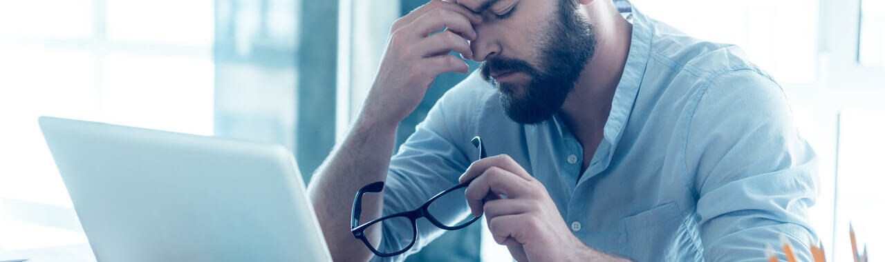 um homem aparentemente cansado, na frente de um computador, representando os sintomas da Síndrome de Burnout
