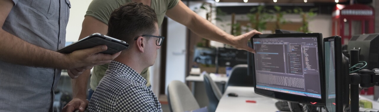 Nesta imagem, podemos observar três pessoas em ação: uma delas está sentada de frente para a tela do computador, enquanto outra pessoa está ao seu lado esquerdo, explicando algo e tocando o monitor. A terceira pessoa está atrás delas, segurando um tablet. A cena simboliza uma solução de treinamento e desenvolvimento.