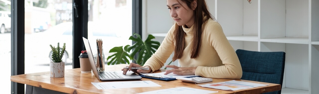 Mulher asiática trabalhando em escritório utilizando computador em mesa corporativa repleta de papéis. 