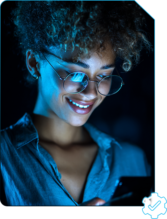 Mulher negra de cabelo cacheado usando óculos redondo e camiseta azul, em ambiente escuro e com luz azul, olhando para o celular e sorrindo